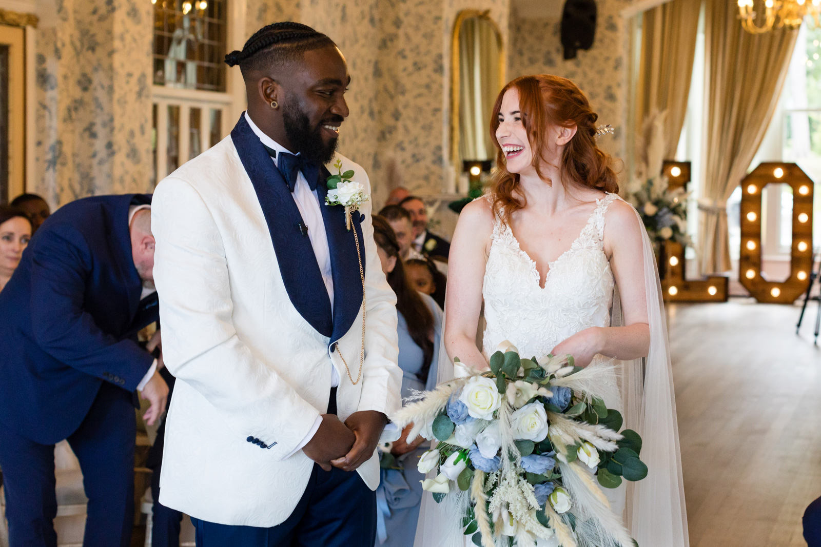 White bride with long red hair smiles and laughs with black groom wearing a white and blue tuxedo at Rushpool Hall wedding