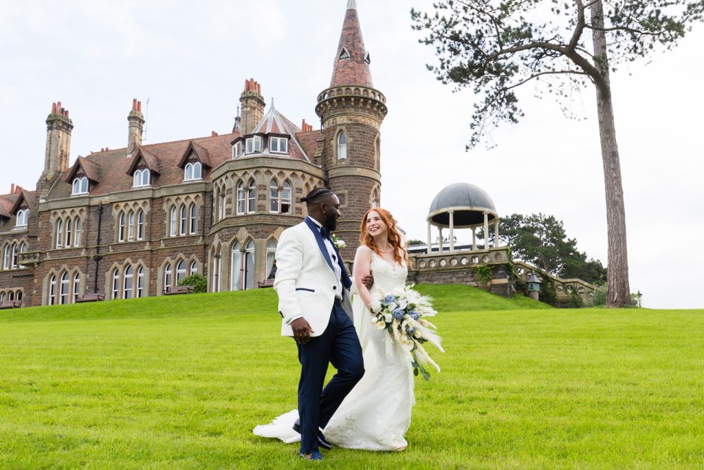 Black groom in white and blue tuxedo walks next to redheaded bride at Rushpool Hall wedding with the building in the background
